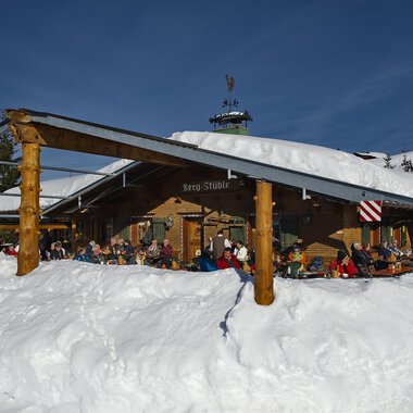 A cozy mountain cabin in the snow with many people on the terrace. The surroundings are wintry and the sun is shining in the blue sky. | © Bergstüble | Michaela Beck Dornach