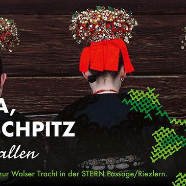 An exhibition about Walser attire featuring three individuals wearing traditional clothing and intricately designed headgear. The background is wood-colored and the colors of the clothing are striking.