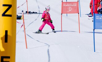 A small skier in pink ski gear is skiing through a gate on a snowy slope. In the background, other skiers and color markings can be seen. | © Skischule Hirschegg | Clemens Paul