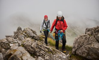 Two mountaineers are moving along a rocky slope. It is foggy, and the surroundings appear wild and untouched. | © Bergschule Kleinwalsertal | Oliver Farys