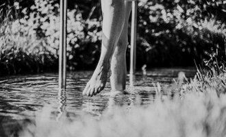 A foot stands in the water, surrounded by green grass. The shot is in black and white and conveys a calm atmosphere. | © Das Naturhotel Chesa Valisa****s