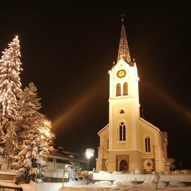 Eine verschneite Landschaft mit einer beleuchteten Kirche und einem geschmückten Weihnachtsbaum. Der Nachthimmel verstärkt die festliche Atmosphäre. | © Kleinwalsertal Tourismus | Frank Drechsel