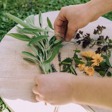 Eine Hand arrangiert frische Kräuter und Blumen auf einem Holzbrett. Der Hintergrund besteht aus grünem Gras. | © Das Naturhotel Chesa Valisa****s