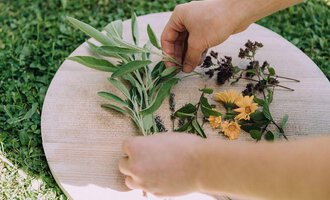 Eine Hand arrangiert frische Kräuter und Blumen auf einem Holzbrett. Der Hintergrund besteht aus grünem Gras. | © Das Naturhotel Chesa Valisa****s