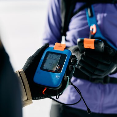 A person is holding a transceiver device in their hand. In the background, a snowy area is visible. | © Kleinwalsertal Tourismus | Oliver Farys
