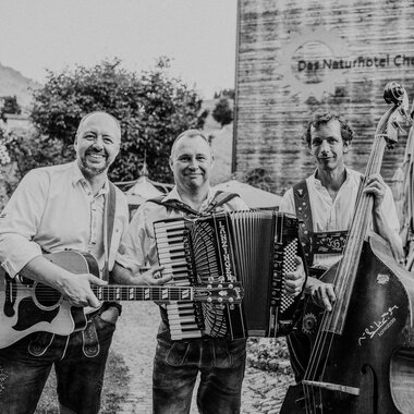 Three musicians are happily gathered together, playing various instruments. In the background, the nature hotel can be seen. | © Naturhotel Chesa Valisa