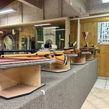 A series of sporting rifles on display stands in a shooting range. In the background, instructions and information about shooting sports can be seen. | © Luftgewehrstand | Michael Veit