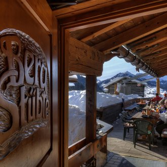 A cozy inn with wooden decorations and a view of snow-covered mountains. Outside, guests are sitting at a table and enjoying the alpine atmosphere. | © Bergstüble | Michaela Beck Dornach