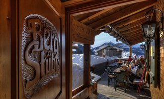 A cozy inn with wooden decorations and a view of snow-covered mountains. Outside, guests are sitting at a table and enjoying the alpine atmosphere. | © Bergstüble | Michaela Beck Dornach