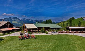 A cozy farmhouse with a large green lawn and a view of the mountains. In the foreground, there is a tractor and there are some outdoor seating options. | © Bergstüble | Michaela Beck