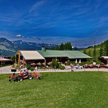 A cozy farmhouse with a large green lawn and a view of the mountains. In the foreground, there is a tractor and there are some outdoor seating options. | © Bergstüble | Michaela Beck