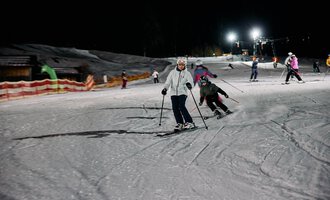 Skiers are waiting at an illuminated ski lift at night. The slopes are covered in snow and surrounded by trees. | © Kleinwalsertal Tourismus | Oliver Farys