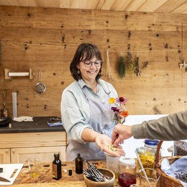 Eine lächelnde Frau in einer Holzküche reicht einer Person Blumen und kleine Flaschen. Im Hintergrund sind Kräuter und verschiedene Zutaten zu sehen. | © Kleinwalsertal Tourismus - Bettina Kessler