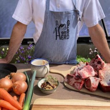 Ein Koch steht an einem Tisch mit frischen Zutaten, darunter Gemüse und Fleisch. Im Hintergrund sind Blumen und eine helle Umgebung sichtbar. | © Gerd am Herd | Gerd Hammerer