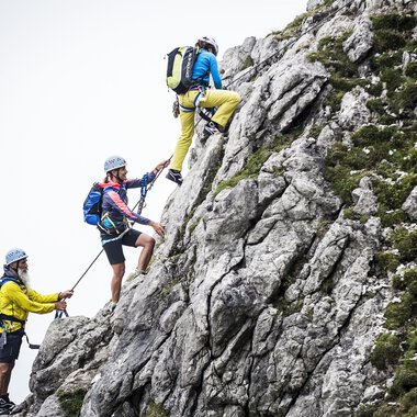 Three climbers are ascending a steep rock. They are wearing helmets and climbing gear as they securely hold on to ropes. | © Bergschule Kleinwalsertal | Dominik Berchtold