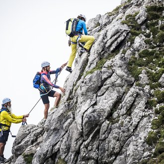 Drei Kletterer erklimmen einen steilen Felsen. Sie tragen Helme und Kletterausrüstung, während sie sich sicher an Seilen festhalten. | © Bergschule Kleinwalsertal | Dominik Berchtold