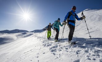 A group of people is hiking through a snowy landscape. The sun is shining clearly in the sky. | © Kleinwalsertal Tourismus eGen | Dominik Berchtold
