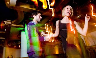 A couple dances happily in a lively club. Colorful lights and a festive atmosphere surround them. | © Aparthotel Kleinwalsertal Haller GmbH & Co KG