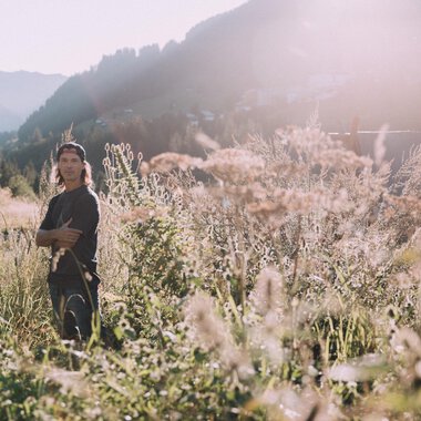 Ein junger Mann steht in einem blühenden Feld mit hohen Gräsern. Die Sonne scheint und die Berge sind im Hintergrund sichtbar. | © Das Naturhotel Chesa Valisa****s