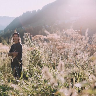 A young man stands in a blooming field with tall grasses. The sun is shining and the mountains are visible in the background. | © Das Naturhotel Chesa Valisa****s