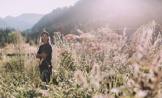 A young man stands in a blooming field with tall grasses. The sun is shining and the mountains are visible in the background. | © Das Naturhotel Chesa Valisa****s