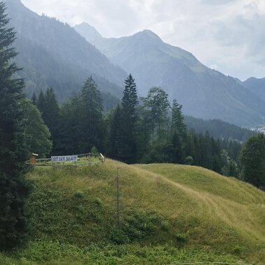 Eine malerische Landschaft mit grünen Hügeln und hohen Bergen. Im Vordergrund steht eine kleine Hütte umgeben von Bäumen. | © Nicole Lughammer