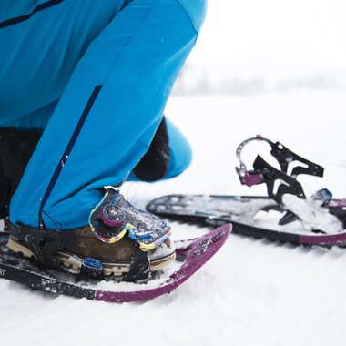 A person straps on snowshoes to hike in the snow. The surroundings are wintry and covered in snow. | © Kleinwalsertal Tourismus | Oliver Farys