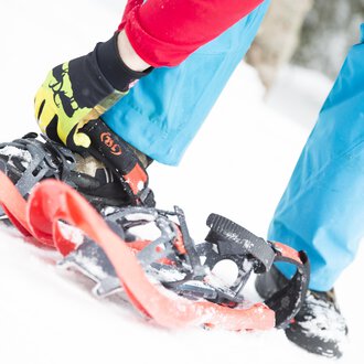 A person is fastening snowshoes while standing on snow. The person is wearing a red jacket and blue pants, creating a colorful contrast to the wintry surroundings. | © Kleinwalsertal Tourismus eGen | Oliver Farys