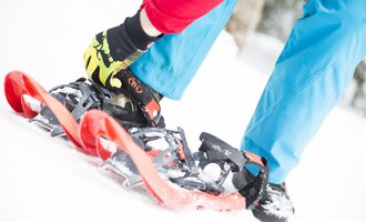 A person is fastening snowshoes while standing on snow. The person is wearing a red jacket and blue pants, creating a colorful contrast to the wintry surroundings. | © Kleinwalsertal Tourismus eGen | Oliver Farys