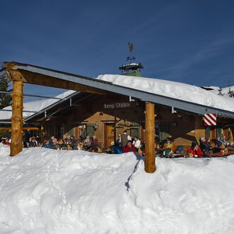 A cozy mountain cabin in the snow with guests on the terrace. The sky is clear and blue, and the surroundings are wintry. | © Bergstüble | Michaela Beck Dornach
