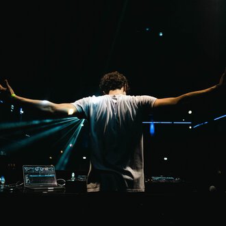 A DJ stands behind his mixing console with outstretched arms. Colorful lights shine in the background, creating a festive atmosphere. | © dj let´s fetz