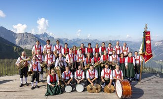 A brass band in traditional attire stands on a platform in the mountains. Impressive mountains and a blue sky are visible in the background. | © Trachtenkapelle Riezlern
