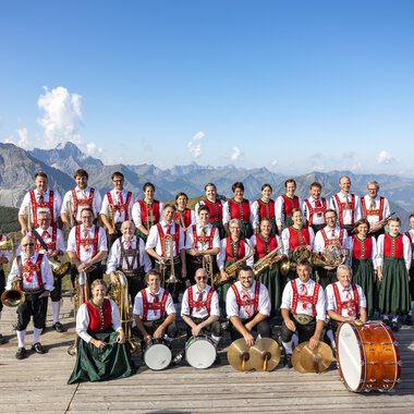 A brass band in traditional attire stands on a platform in the mountains. Impressive mountains and a blue sky are visible in the background. | © Trachtenkapelle Riezlern