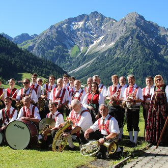 © Musikverein d´Hirschegger | Jockl Metzger Eine Musikkapelle in traditioneller Tracht steht vor einer malerischen Berglandschaft. Im Hintergrund sind grüne Wiesen und schneebedeckte Berge zu sehen. | © Musikverein d´Hirschegger | Jockl Metzger