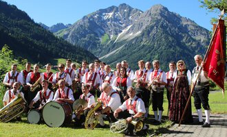 Eine Musikkapelle in traditioneller Tracht steht vor einer malerischen Berglandschaft. Im Hintergrund sind grüne Wiesen und schneebedeckte Berge zu sehen. | © Musikverein d´Hirschegger | Jockl Metzger