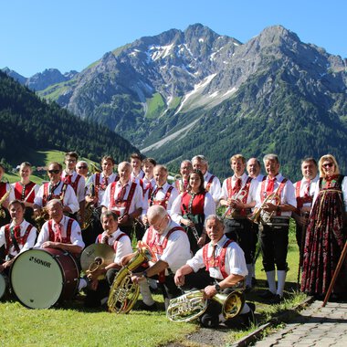 Eine Musikkapelle in traditioneller Tracht steht vor einer malerischen Berglandschaft. Im Hintergrund sind grüne Wiesen und schneebedeckte Berge zu sehen. | © Musikverein d´Hirschegger | Jockl Metzger