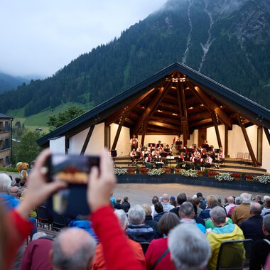 Eine Freiluftbühne in den Bergen mit einer Orchesteraufführung. Viele Zuschauer sitzen davor und genießen die Musik in der Abenddämmerung. | © Kleinwalsertal Tourismus | Oliver Farys