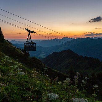 © OBERSTDORF · KLEINWALSERTAL BERGBAHNEN Eine Seilbahn fährt über hügelige Berge bei Sonnenuntergang. Der Himmel ist in warmen Farben erleuchtet. | © OBERSTDORF · KLEINWALSERTAL BERGBAHNEN
