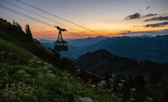 Eine Seilbahn fährt über hügelige Berge bei Sonnenuntergang. Der Himmel ist in warmen Farben erleuchtet. | © OBERSTDORF · KLEINWALSERTAL BERGBAHNEN
