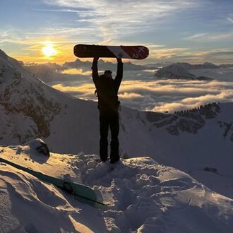 A snow sports enthusiast stands on a snowy peak, holding a snowboard up high. In the background, the sun shines between the mountains and a blanket of clouds glows in golden colors. | © Abenteuer Vertical | Christian Kohler