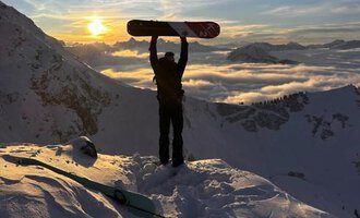 A snow sports enthusiast stands on a snowy peak, holding a snowboard up high. In the background, the sun shines between the mountains and a blanket of clouds glows in golden colors. | © Abenteuer Vertical | Christian Kohler