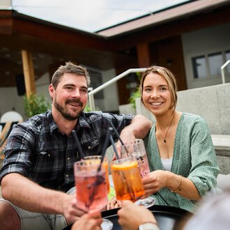 Ein fröhliches Paar, das zusammen Cocktails genießt. Im Hintergrund sind gemütliche Sitzgelegenheiten sichtbar. | © Kleinwalsertal Tourismus | Oliver Farys
