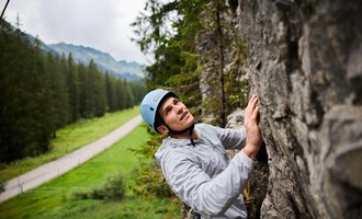 A climber working on a rock face is wearing a helmet and concentrating on his ascent. In the background, trees and a road can be seen in a green landscape. | © Bergschule Kleinwalsertal | Oliver Farys