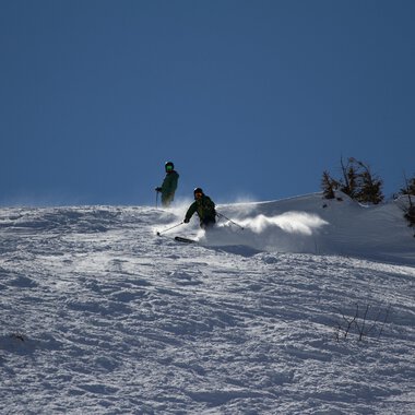 Two skiers are going down a snow-covered slope. The sky is clear and blue. | © Markus Hoefler