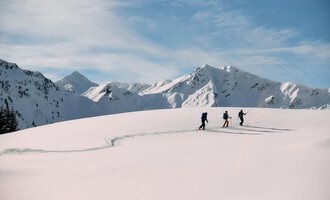 A skier in a blue suit is skiing through fresh, powdery snow. In the background, snow-covered mountains and a clear blue sky can be seen. | © Kleinwalsertal Tourismus | Oliver Farys
