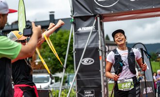 Eine Triathletin läuft glücklich über die Ziellinie und wird von ihren Unterstützern gefeiert. Im Hintergrund sind die Veranstaltungstafeln und eine grüne Landschaft zu sehen. | © Kleinwalsertal Tourismus - Dominik Berchtold