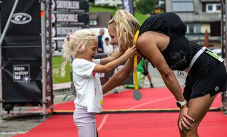 Ein fröhliches Mädchen überreicht einer Sportlerin eine Medaille nach einem Wettkampf. Die Atmosphäre ist von Freude und Stolz geprägt. | © Kleinwalsertal Tourismus - Dominik Berchtold