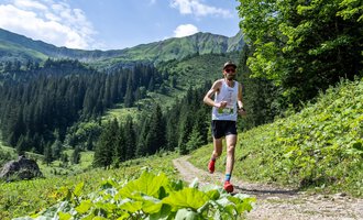 Ein Läufer joggt auf einem schmalen Weg in den Bergen. Die Landschaft ist grün mit herrlichen Wäldern und hohen Bergen im Hintergrund. | © Kleinwalsertal Tourismus - Dominik Berchtold