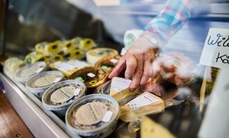 A sales stand with various types of cheese. A hand reaches for a cheese product in the display. | © Kleinwalsertal Tourismus | Fotograf Oliver Farys