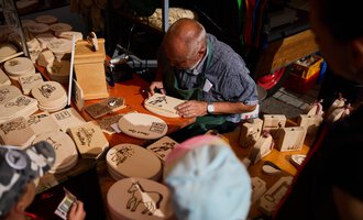 An older man is carving wooden figures at a market stand. Around him lie numerous wooden boards with various motifs. | © Kleinwalsertal Tourismus | Fotograf Oliver Farys